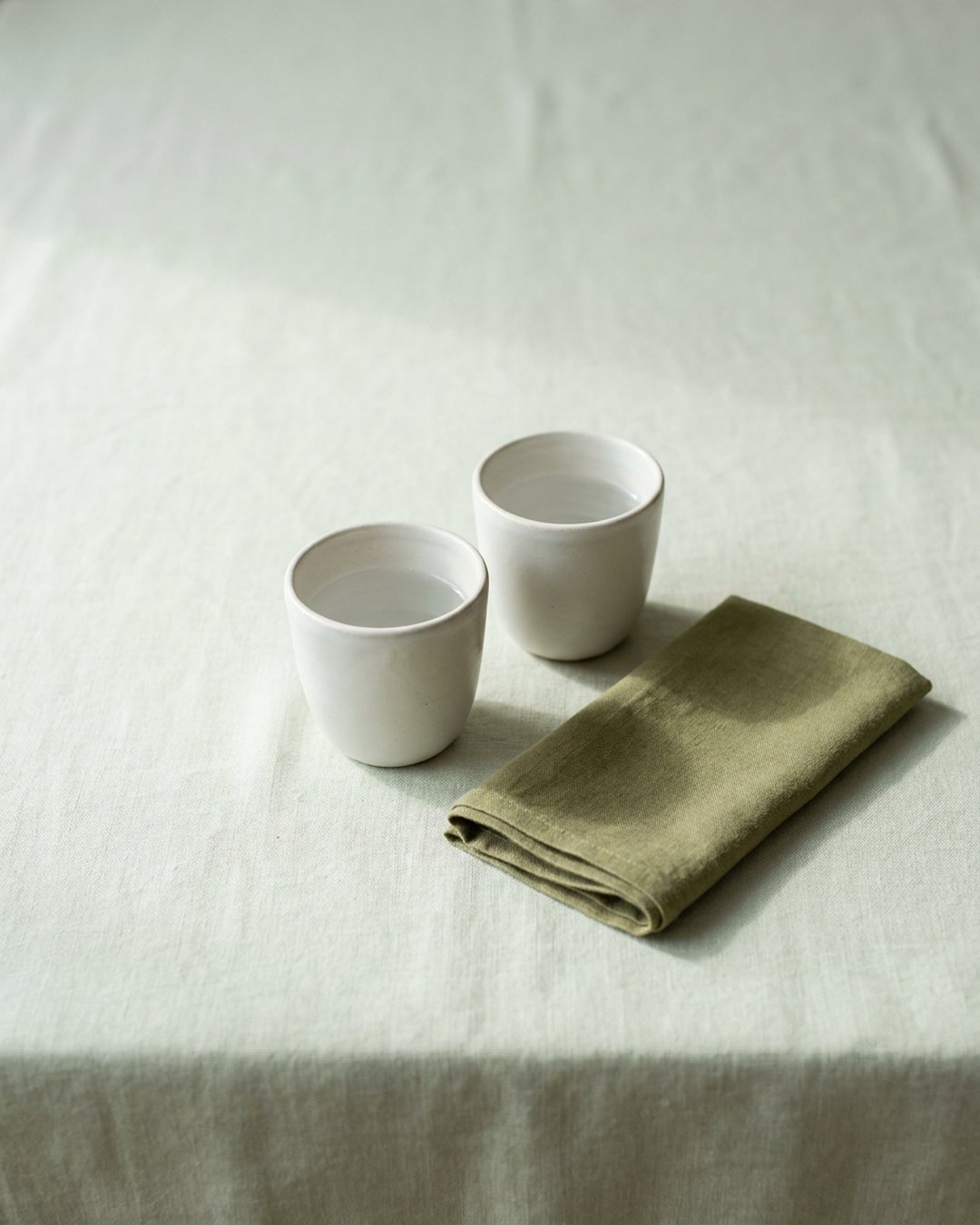 Two small white ceramic cups of water side by side on a sage linen tablecloth